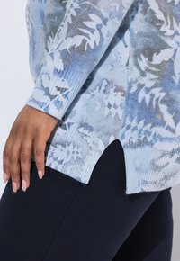 Maglione azzurro chiaro con un motivo a foglia, polsini a costine e spacchi laterali. Indossato con leggings neri, evidenziando la texture e il design.