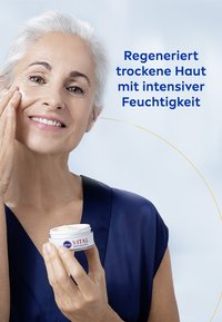 White cream in a blue and white jar labeled "NIVEA VITAL ANTI-FALTEN" held by a person applying cream to their face, wearing a dark blue top.