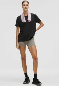 Sort kortærmet t-shirt, beige tætsiddende shorts, lyserødt håndklæde draperet om halsen, sorte sneakers og sorte sokker. Simpel atletisk design.