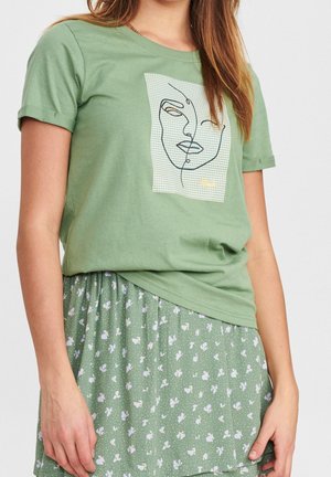 Femme portant un t-shirt vert avec un dessin abstrait de visage et une jupe florale verte, debout devant un fond blanc uni.