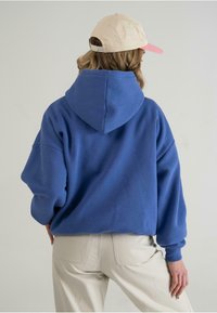 Blå oversized hoodie lavet af blødt stof med snor i hætten og manchetter, parret med lyse beige bukser og en tan kasket.