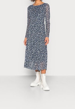 Frau trägt ein marineblaues Midikleid mit weißen und beigen Punkten und langen Ärmeln, kombiniert mit weißen klobigen Stiefeletten.