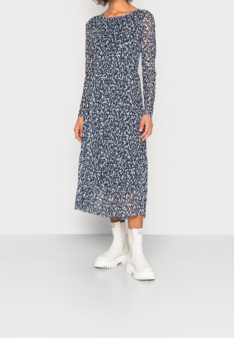 Femme portant une robe midi bleu marine avec des taches blanches et beiges et des manches longues, associée à des bottines blanches épaisses.