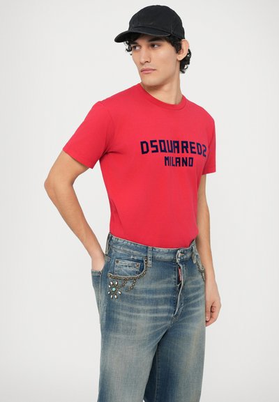 Červené bavlnené tričko s nápisom „DSQUARED2 MILANO“ v námorníckej modrej potlači, v páre s modrými rifľami so zdobnými cvočkami a jedinečným dizajnom vrecka.