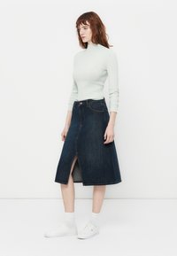 Jeans A-linje skjørt med en frontslitt, mørkeblå farge, kombinert med en lys grønn ribbestrikket topp og hvite joggesko, stående mot en hvit bakgrunn.