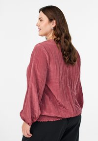 Žena s valovitom smeškom kosom koja nosi srebrne naušnice, teksturiranu roza bluzu s dugim rukavima i crne hlače, prikazana sa stražnje i bočne strane.