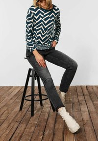Maglione a maniche lunghe in blu scuro con motivo a zigzag bianco, abbinato a jeans neri sbiaditi e stivaletti cremisi, seduto su uno sgabello nero.