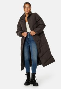 Lang, mørkebrun dunjakke med høj krave, quiltet design, sidelommer og hel lynlås. Bæres over tætsiddende jeans og støvler.