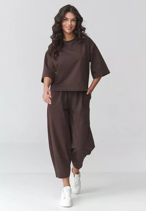 Kobieta idąca w luźnej, ciemnobrązowej bluzce i szerokich spodniach, w białych trampkach, na jasnym, jednolitym tle.