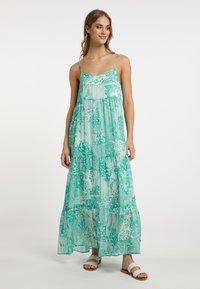 Rochie maxi cu imprimeu floral verde, cu bretele subțiri, design în straturi și material ușor, semi-transparent. Sandale plate completează aspectul.