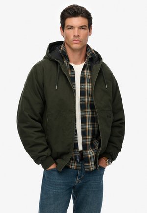 Green hooded jacket with a matte finish worn over a blue and green plaid shirt, paired with blue jeans. Features front pockets and drawstrings.