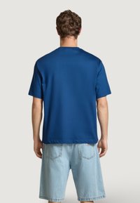 Muž oblečený v jednoduchém modrém tričku s krátkým rukávem a světlých džínových šortkách, stojící zády k fotoaparátu proti světlému pozadí.