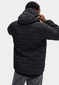 Giacca a vento nera con cappuccio, caratterizzata da un design trapuntato e un tessuto testurizzato. Il modello sta regolando il cappuccio.