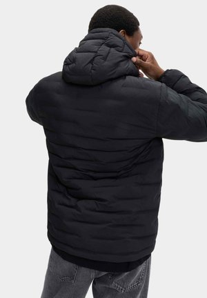 Schwarze Daunenjacke mit Kapuze, die ein gesteppter Entwurf und strukturierten Stoff aufweist. Das Modell justiert die Kapuze.