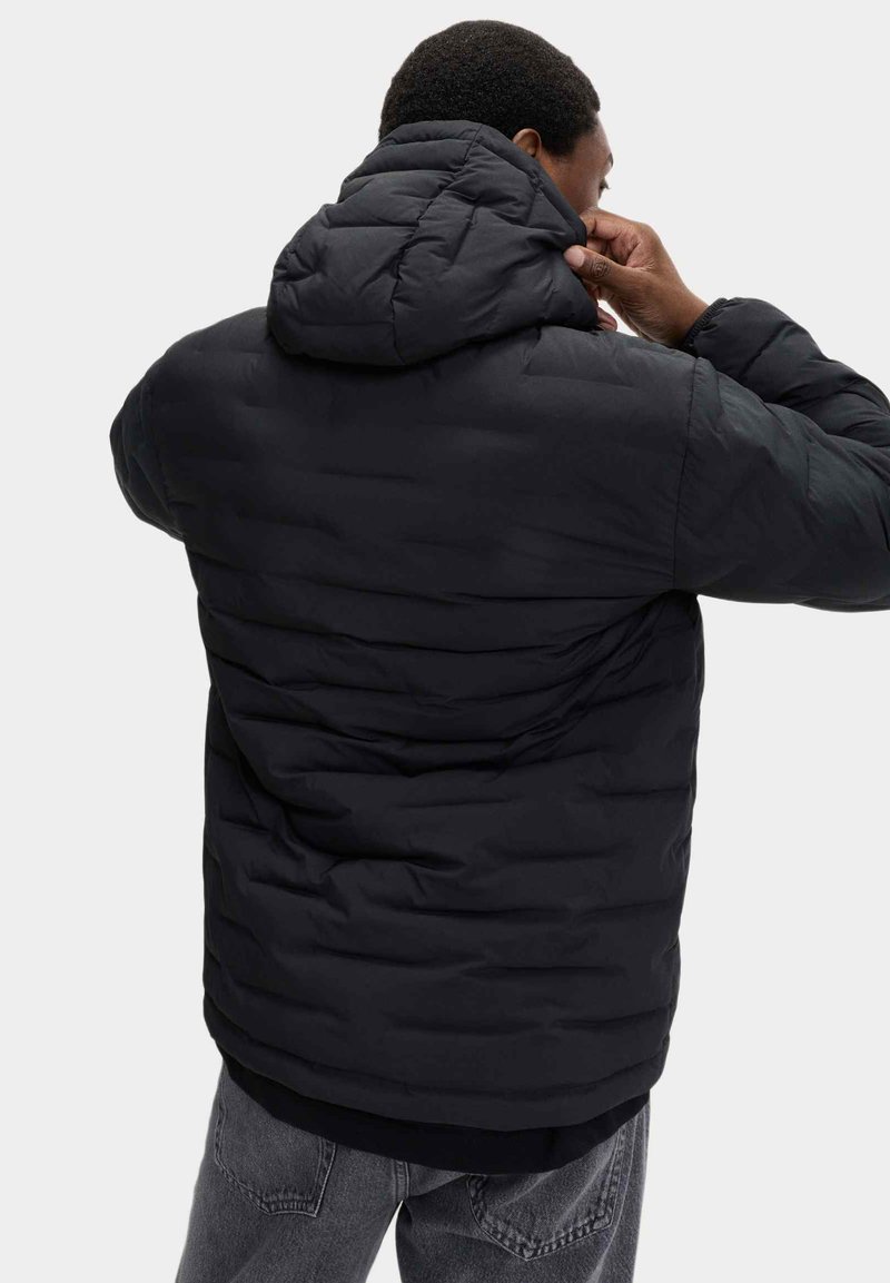 Giacca a vento nera con cappuccio, caratterizzata da un design trapuntato e un tessuto testurizzato. Il modello sta regolando il cappuccio.