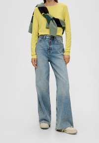 Gelbes Langarmshirt, kombiniert mit hoch taillierten, weit geschnittenen blauen Jeans. Ein grün-schwarz gestreiftes Sweatshirt über die Schultern gelegt.