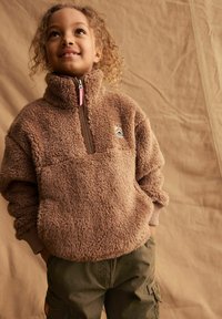 Pullover in pile marrone, morbido, con zip, colletto alto e polsini a coste; presenta un piccolo logo colorato sul petto; abbinato a pantaloni cargo verdi.