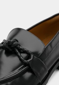Μαύρο δερμάτινο μοκασίνι με διακοσμητικό πλεγμένο κόμπο, λεία υφή, στρογγυλεμένη μύτη και εσωτερική επένδυση σε ζεστή καφέ απόχρωση.