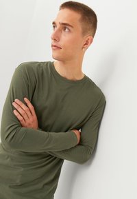 Langsleeve, olijfgroen T-shirt van zachte stof. Heeft een ronde halslijn en aansluitende mouwen. Model staat met gekruiste armen.