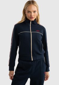 Veste de survêtement zippée marine avec col haut, bandes blanches et rouges contrastantes le long des manches, et logo brodé sur la poitrine. Tissu doux et texturé.