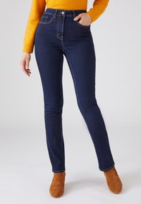 Jeans de talle alto de color azul oscuro con costuras sutiles, que presentan bolsillos frontales y un diseño ajustado, combinados con botines de ante marrón.