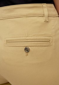 Pantalons beiges avec une poche arrière à ouverture horizontale et fermeture à bouton circulaire, fabriqués en tissu lisse avec des détails de couture.