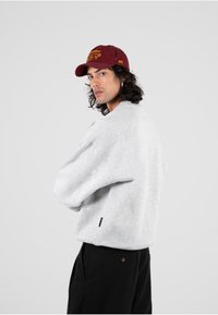 Felpa grigia in una vestibilità rilassata, con scollo rotondo e una texture sottile. Abbinata a pantaloni neri e un berretto bordeaux con testo giallo.