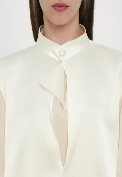 Cream-colored blouse in smooth satin with a stand collar, featuring a flap detail, a single button closure, and sheer accents.
