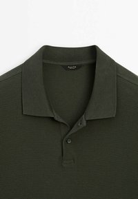 Polo vert foncé texturé avec un col plié et deux boutons au niveau de l'encolure, posé à plat sur un fond blanc.