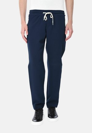 Uomo con pantaloni tuta blu navy con cordino bianco e sneakers nere con suola bianca, in piedi davanti a uno sfondo neutro.