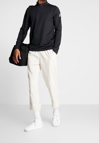 Schwarzer Pullover mit langen Ärmeln und Stehkragen, kombiniert mit cremefarbenen schmalen Hosen und weißen Sportschuhen, eine schwarze Tasche haltend.