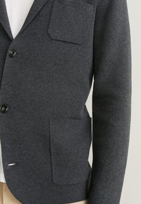 Cardigan in lana grigia scura con bottoni e due tasche frontali, indossato sopra una camicia bianca e pantaloni beige, che mostra la parte centrale del torso e il braccio destro.