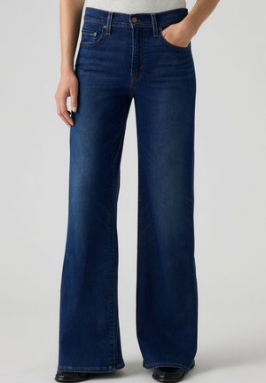 Mørkeblå, højttaljede, vide jeans båret med den ene hånd i lommen og sorte sko synlige ved kanten.