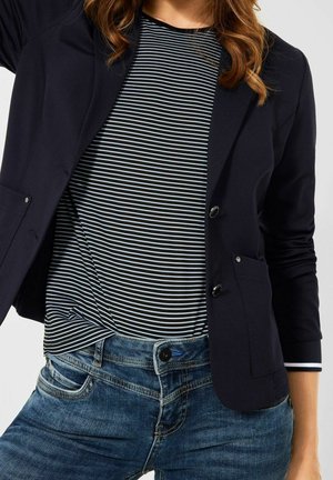 Kobieta w czarnym blezerze, czarno-białej koszuli w paski i niebieskich jeansach na gładkim tle.