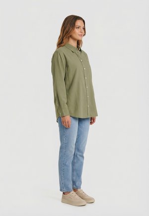 Donna in piedi di lato che indossa una camicia verde oliva oversize con bottoni, jeans azzurri e sneakers beige su sfondo grigio uniforme.