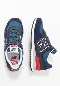 Pár modro-červených tenisek New Balance 574 s prodyšnou síťovinou a semišovým povrchem na bílém pozadí.