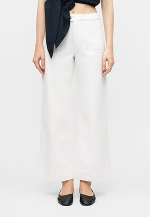 Nő magas derekú, széles szárú, fehér nadrágot visel elülső zsebekkel, fekete rövid felsővel és fekete lapos cipővel párosítva.