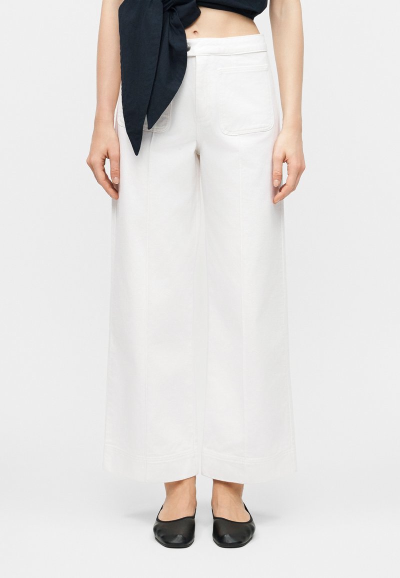 Γυναίκα που φοράει λευκό παντελόνι ψηλόμεσο με φαρδιά πόδια και μπροστινές τσέπες, σε συνδυασμό με μαύρο κοντό τοπ και μαύρα επίπεδα παπούτσια.