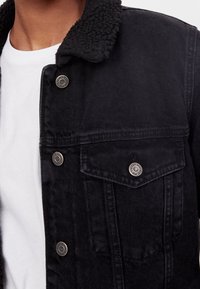 Svart denimjacka med en fluffig svart krage, framknappar och bröstficka. Har en strukturerad tyg och en ren, modern design.