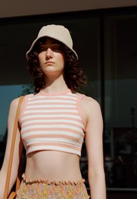Top cropped com riscas em rosa e branco, combinado com uma saia de folhos multicolorida. Apresenta um chapéu tipo balde bege e uma mala de ombro castanha.