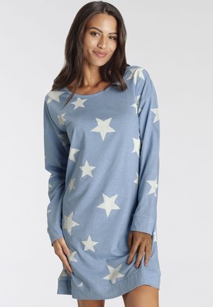 Frau trägt ein blaues Langarmkleid mit weißen Sternmustern, steht mit den Händen nahe den Oberschenkeln und lächelt leicht vor einem einfarbigen Hintergrund.