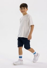 T-shirt beige chiaro a maniche corte con un piccolo etichetta; pantaloni cargo scuri; scarpe da ginnastica bianche con calzini a strisce blu; sfondo neutro.