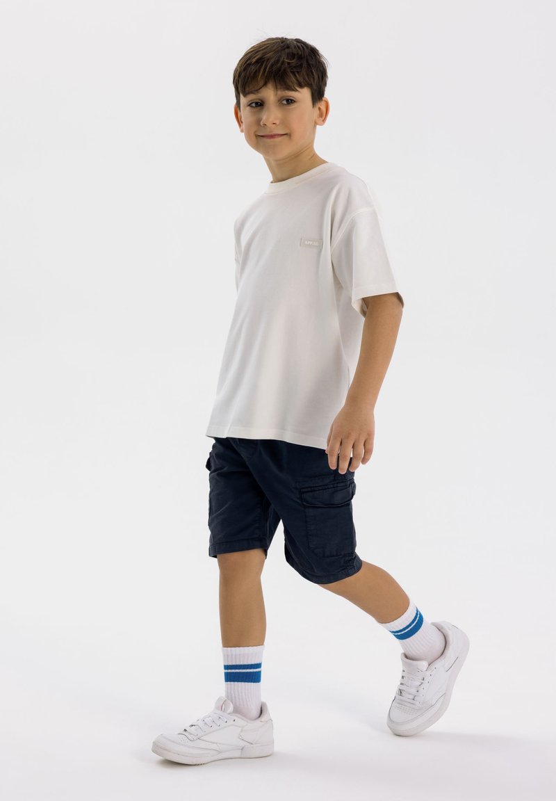 T-shirt beige chiaro a maniche corte con un piccolo etichetta; pantaloni cargo scuri; scarpe da ginnastica bianche con calzini a strisce blu; sfondo neutro.