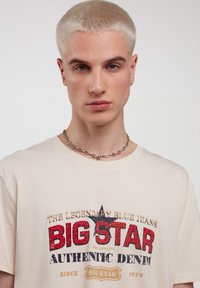Kreminės spalvos marškinėliai su raudonos, mėlynos ir auksinės spalvos grafiką, featuring tekstą „BIG STAR AUTHENTIC DENIM“ ir žvaigždės dizainą. Sidabrinė grandinėlė.