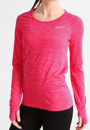 Femme portant un haut d'entraînement Nike à manches longues, rose vif, avec des trous pour les pouces, associé à des leggings noirs.