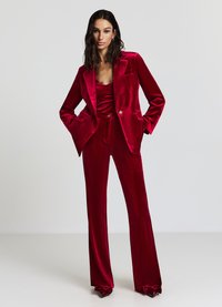 Costume en velours rouge comprenant un blazer ajusté à revers et un pantalon à jambes larges. Les accents incluent un bouton doré et un détail de nœud papillon sur les chaussures.