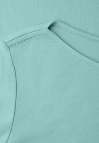 Primer plano de tela de color verde azulado suave que muestra costuras detalladas y líneas de unión de una prenda de algodón, probablemente el área del hombro de una camiseta o sudadera.