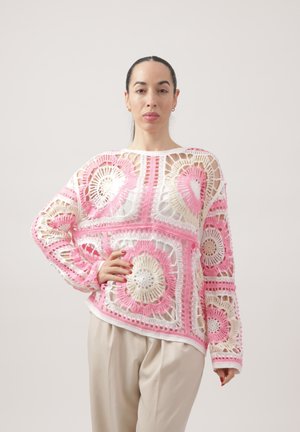 Kobieta z ciemnymi włosami związanymi w kok, ubrana w różowy, biały i beżowy sweter na szydełku oraz beżowe spodnie na jednolitym jasnym tle.