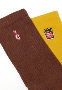 Calcetines marrones y amarillos con textura acanalada; el calcetín marrón presenta un bordado de una botella de ketchup, mientras que el calcetín amarillo tiene un diseño de papas fritas.