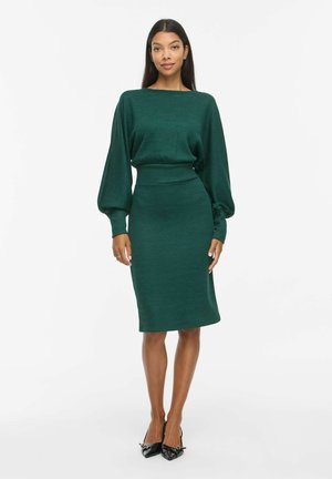 Un mannequin se tient devant un fond blanc, vêtu d’une robe vert foncé à longues manches bouffantes, avec une jupe ajustée au genou et des chaussures plates noires.
